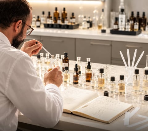 Male perfumer in white shirt examining fragrance samples at table with notebook and multiple bottles in laboratory setting